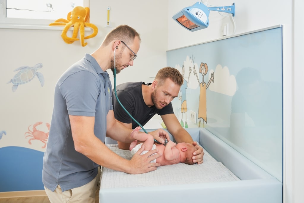 Dr. Christoph Rodler untersucht ein Neugeborenes mit dem Stethoskop während sich der Vater des Neugeborenen lieb darüber beugt