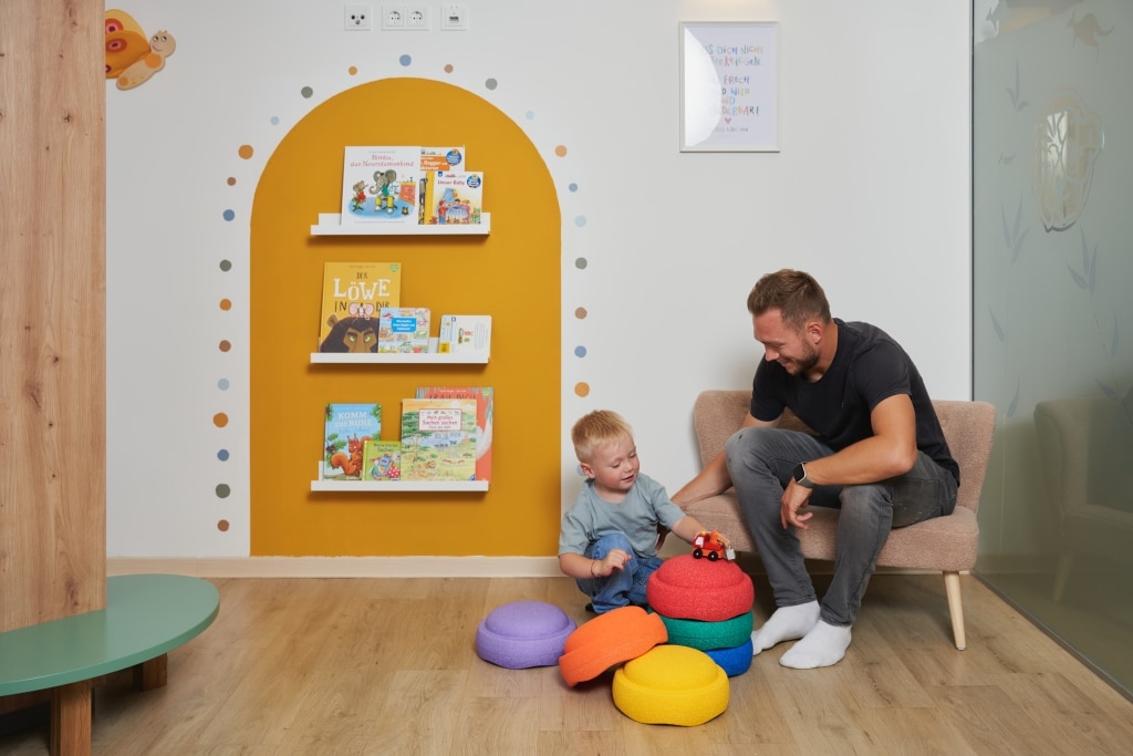 Ein Vater sitzt mit seinem spielenden Sohn im Wartebereich bei Stapelsteinen; man sieht das Bücherregal auf gelb umrandeter Farbe mit bunten Punkten drum herum