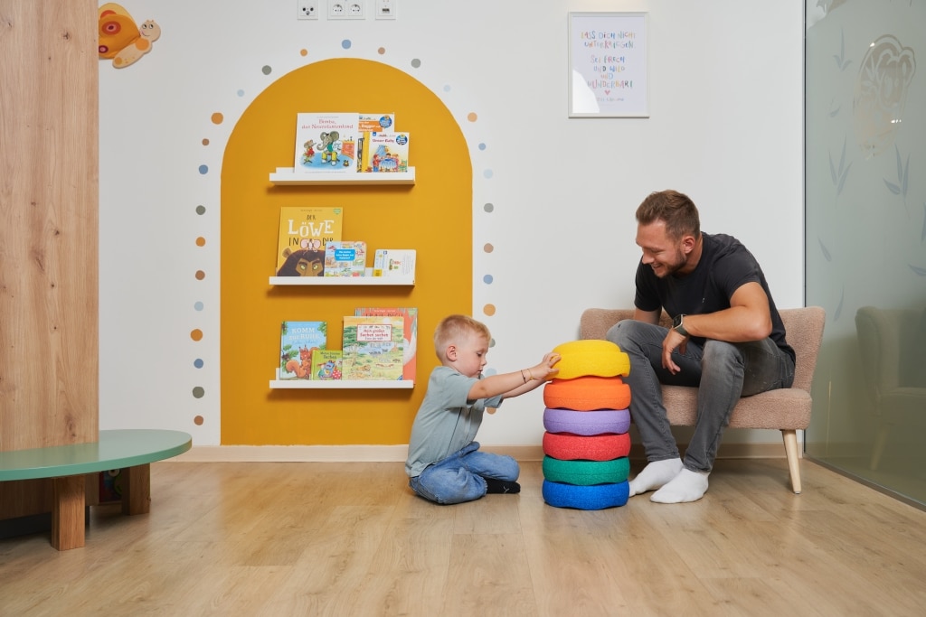 Ein Vater sitzt mit seinem spielenden Sohn im Wartebereich bei Stapelsteinen; man sieht das Bücherregal auf gelb umrandeter Farbe mit bunten Punkten drum herum
